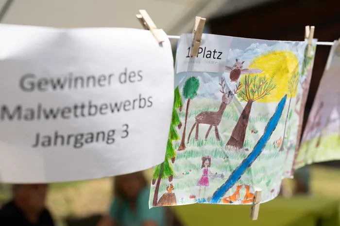 Ein Banner mit der Aufschrift „Gewinner des Malwettbewerbs Jahrgang 3“ hängt neben einem Kinderbild, das einen Hirsch, ein Mädchen und Füchse zeigt. Das Bild ist mit „1. Platz“ und einem Namen beschriftet.