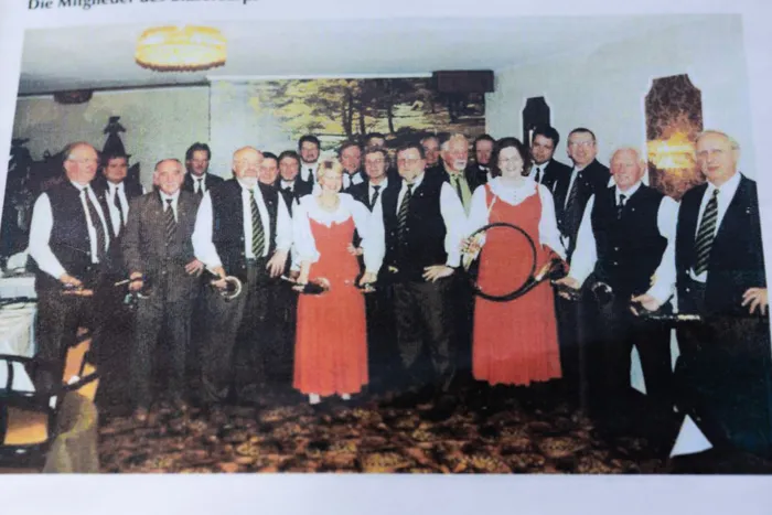 Eine Gruppe von etwa 30 Personen, hauptsächlich Männer in Anzügen, posiert für ein Foto. Im Zentrum stehen zwei Frauen in roten Kleidern mit weißen Oberteilen und halten einen Reifen. Im Hintergrund sind weitere Personen und eine festliche Dekoration zu sehen.