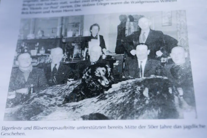 Schwarz-Weiß-Foto von fünf Männern in Anzügen, die um ein erlegtes Wildschwein posieren. Im Hintergrund sind Möbel und ein Fenster zu sehen.