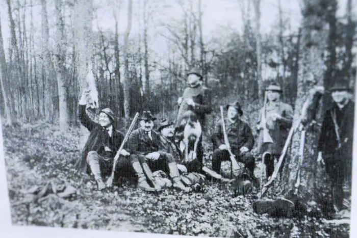 Eine Gruppe von etwa zehn Männern in Jagdkleidung posiert im Wald, einige halten Gewehre oder erbeutete Tiere hoch. Sie feiern das 100-jährige Bestehen des Hegerings.