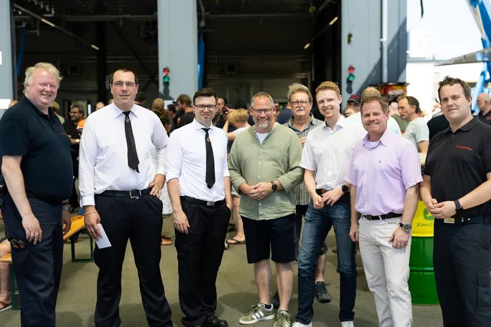 A group of eight men in business or casual attire pose for a photo indoors, with a crowd of people visible in the background. A yellow and green John Deere barrel is visible to the right.