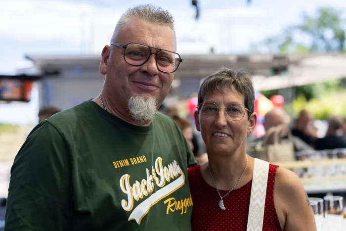Ein Mann mit Bart und Brille und eine Frau mit kurzem Haar lächeln in die Kamera. Im Hintergrund sind Menschen und ein Festzelt zu sehen.