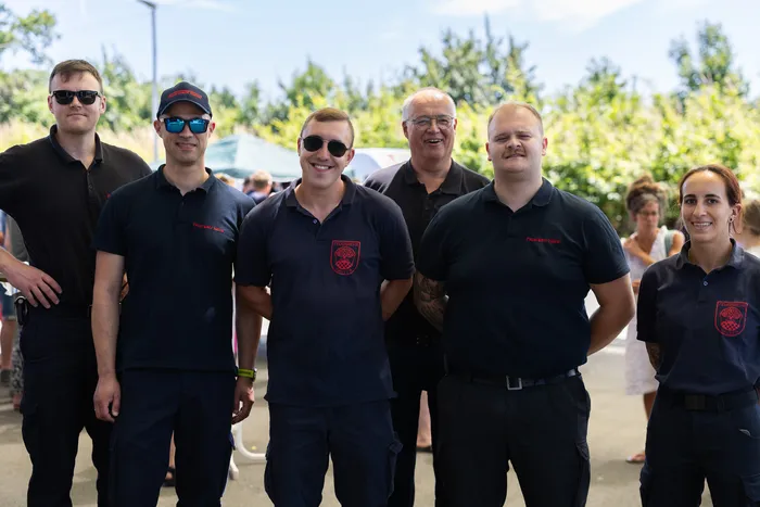 Sechs Personen in dunklen Poloshirts mit Logo stehen lächelnd vor Bäumen und Zuschauern. Die Jubiläumsfeier des Löschzugs Bommert wird im Hintergrund durch Menschen und Festlichkeiten angedeutet.