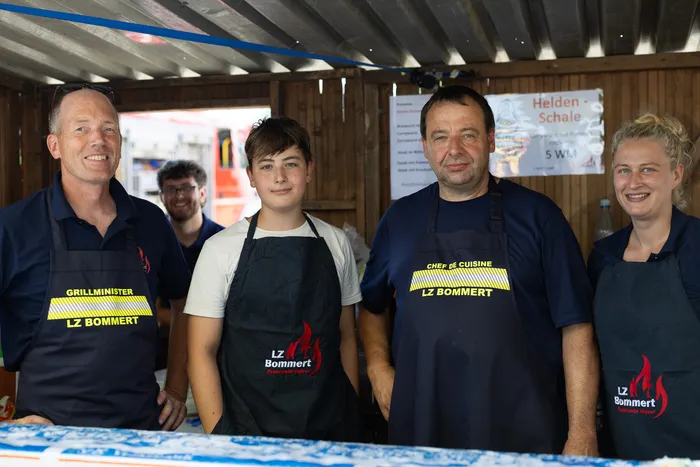 Vier Personen in Schürzen mit dem Aufdruck „LZ Bommert“ stehen vor einem Imbissstand. Links trägt ein Mann ein T-Shirt mit der Aufschrift „Grillminister LZ Bommert“, in der Mitte ein Jugendlicher und rechts eine Frau. Im Hintergrund ist ein Mann zu sehen.