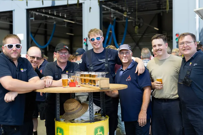 Eine Gruppe von sieben Männern steht lächelnd vor einer offenen Garagentür. Einige halten Biergläser in der Hand, während ein Mann einen Strohhut trägt. Im Hintergrund sind weitere Personen und Fahrzeuge zu sehen.