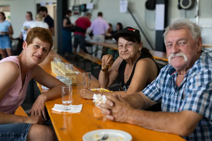 Drei ältere Menschen sitzen an einem Holztisch in einer hellen Halle und essen. Im Hintergrund sind weitere Menschen und Tische zu sehen.