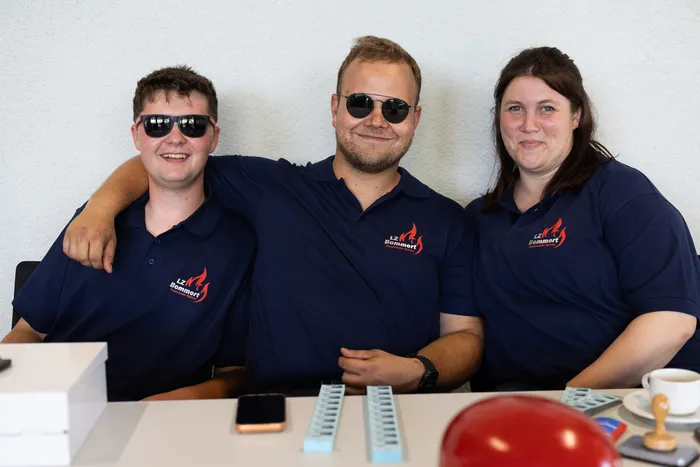 Drei Personen in dunkelblauen Poloshirts mit dem Logo „LZ Bommert“ lächeln in die Kamera. Im Hintergrund steht ein Tisch mit Gegenständen und eine Tasse Kaffee.