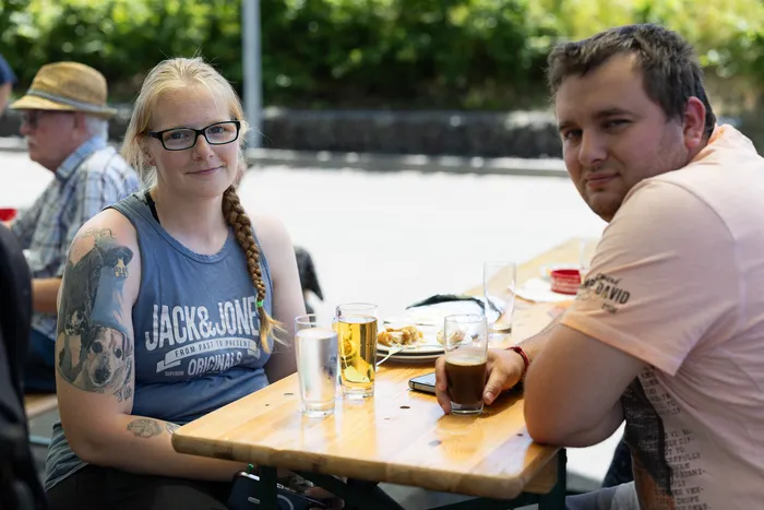Zwei Personen sitzen an einem Holztisch im Freien, vor ihnen Getränke und Snacks. Im Hintergrund sind weitere Personen und ein grüner Garten zu sehen.