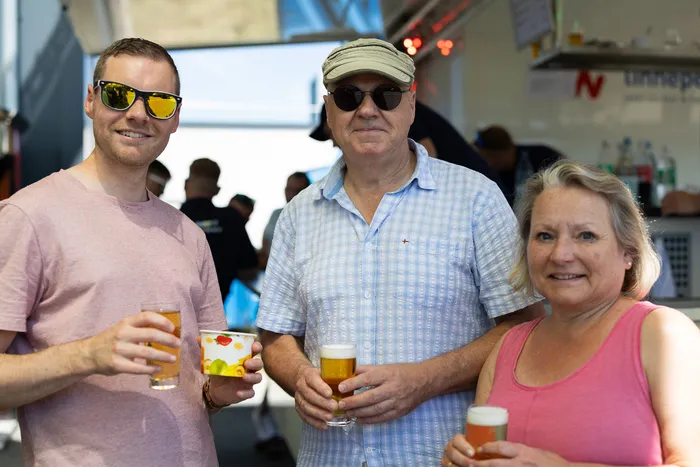Drei Personen – ein junger Mann mit Sonnenbrille, ein älterer Mann mit Kappe und eine Frau – stehen lächelnd mit Getränken in der Hand vor einer Bar. Im Hintergrund sind weitere Menschen zu sehen.