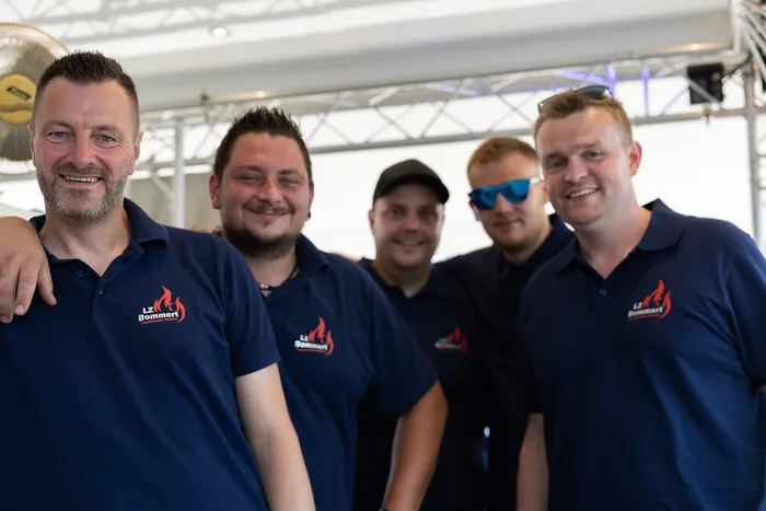 Fünf Feuerwehrleute in dunkelblauen Poloshirts mit dem Logo „LZ Bommert“ lächeln in die Kamera. Im Hintergrund ist ein Zelt und weitere Personen zu sehen.