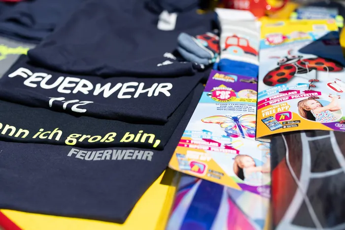 Ein schwarzes T-Shirt mit weißem Aufdruck „FEUERWEHR“ und dem Satz „wenn ich groß bin, FEUERWEHR“ liegt neben bunten Kinderartikeln, darunter ein Pop-up-Buch mit einem Mädchen und ein Marienkäfer-Flugzeug.