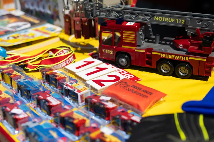 Ein roter Feuerwehrauto-Spielzeugwagen steht neben einem Schild mit der Notrufnummer 112 und weiteren Feuerwehrmemorabilien auf einem Tisch. Im Hintergrund sind weitere Spielzeuge und Artikel zu sehen.