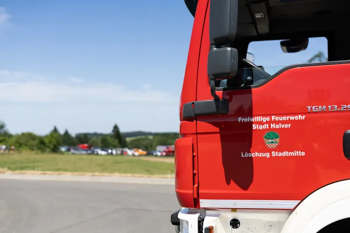 Roter Feuerwehrauto der Freiwilligen Feuerwehr Stadt Halver mit dem Schriftzug 'Löschzug Stadtmitte'. Im Hintergrund verschwommen weitere Fahrzeuge und Landschaft.