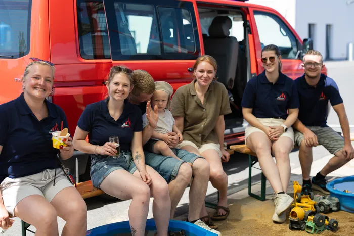 Sechs Feuerwehrleute sitzen auf einer Bank vor einem roten Mannschaftswagen und lächeln in die Kamera. Ein Kind winkt.