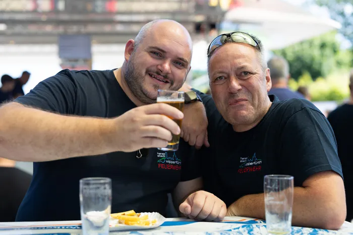 Zwei Männer in T-Shirts der Freiwilligen Feuerwehr stoßen mit Biergläsern an und lächeln in die Kamera. Im Hintergrund sind weitere Personen und ein Gebäude zu sehen.