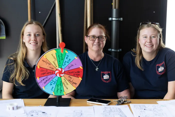 Drei Frauen in dunkelblauen Poloshirts mit dem Logo der Freiwilligen Feuerwehr stehen hinter einem Tisch. In der Mitte steht ein Glücksrad mit verschiedenen Aufgaben für die Feuerwehr.
