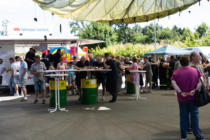 Menschen stehen und unterhalten sich an Stehtischen, die aus grünen Fässern bestehen, vor einem Imbisswagen mit der Aufschrift „W linnepe liefert.“ Im Hintergrund ist eine Hüpfburg und weitere Festlichkeiten zu sehen.