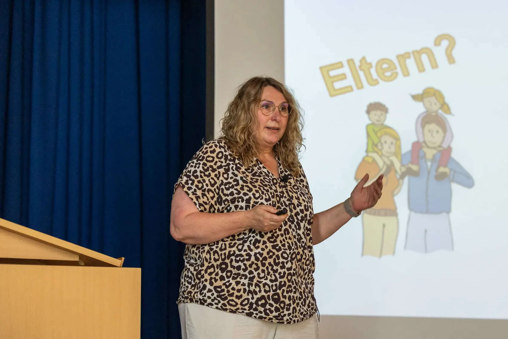 Die Frau mit lockigem Haar spricht vor einem Publikum und gestikuliert dabei. Im Hintergrund ist eine Präsentation mit der Frage „Eltern?“ und stilisierten Figuren von Familien zu sehen.