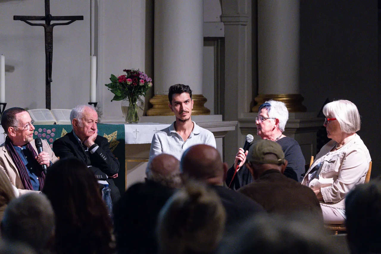 Eine Diskussionsrunde mit mehreren Personen in einer Kirche, einige halten Mikrofone, im Hintergrund sind ein Altar und ein Kruzifix zu sehen.