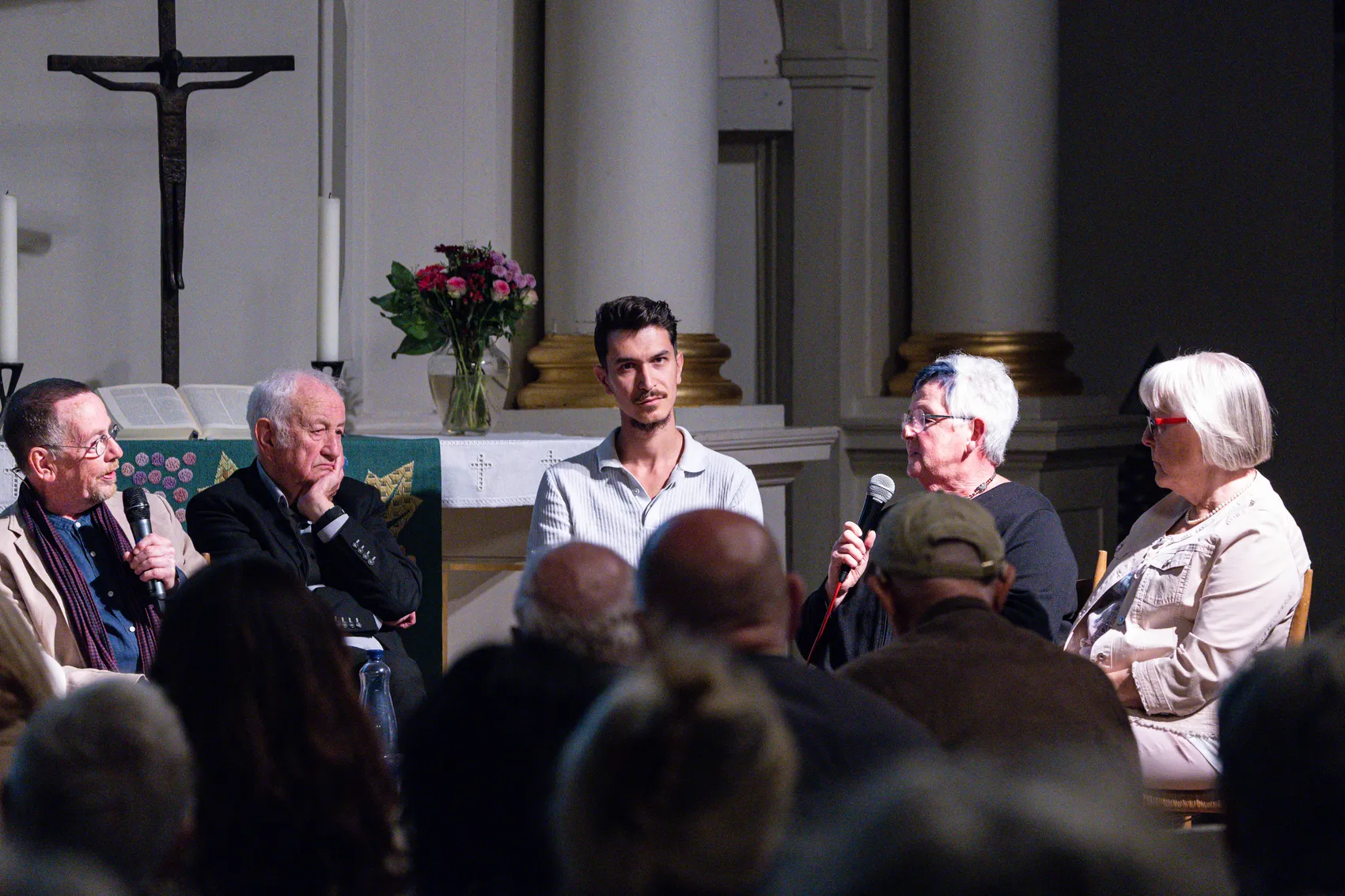 Eine Diskussionsrunde mit mehreren Personen in einer Kirche, einige halten Mikrofone, im Hintergrund sind ein Altar und ein Kruzifix zu sehen.