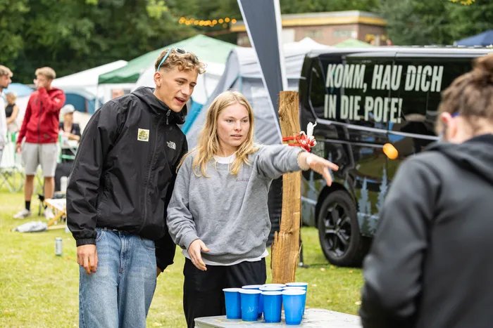Junge Menschen spielen ein Spiel mit blauen Bechern beim Bautz Festival vor einem schwarzen Van mit der Aufschrift „KOMM, HAU DICH IN DIE POFE!“.
