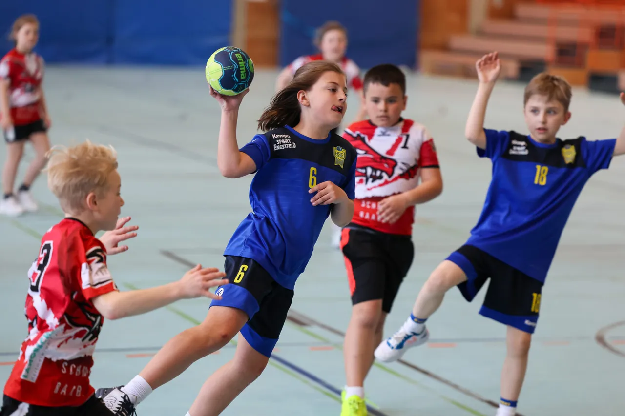 Ein Handballspieler in rotem Trikot mit der Nummer 14 springt beim Wurf, während ein Gegenspieler in blauem Shorts ihn verteidigt. Im Hintergrund sind Zuschauer in einer Sporthalle zu sehen.