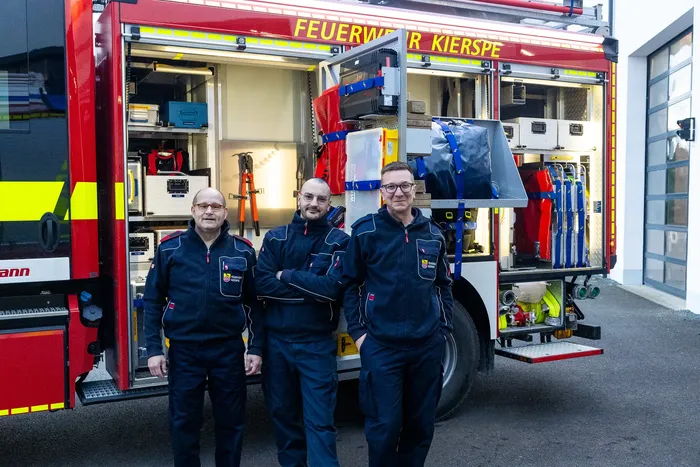 Drei Feuerwehrleute in Uniform stehen vor einem roten Feuerwehrauto mit der Aufschrift „Feuerwehr Kierspe“.