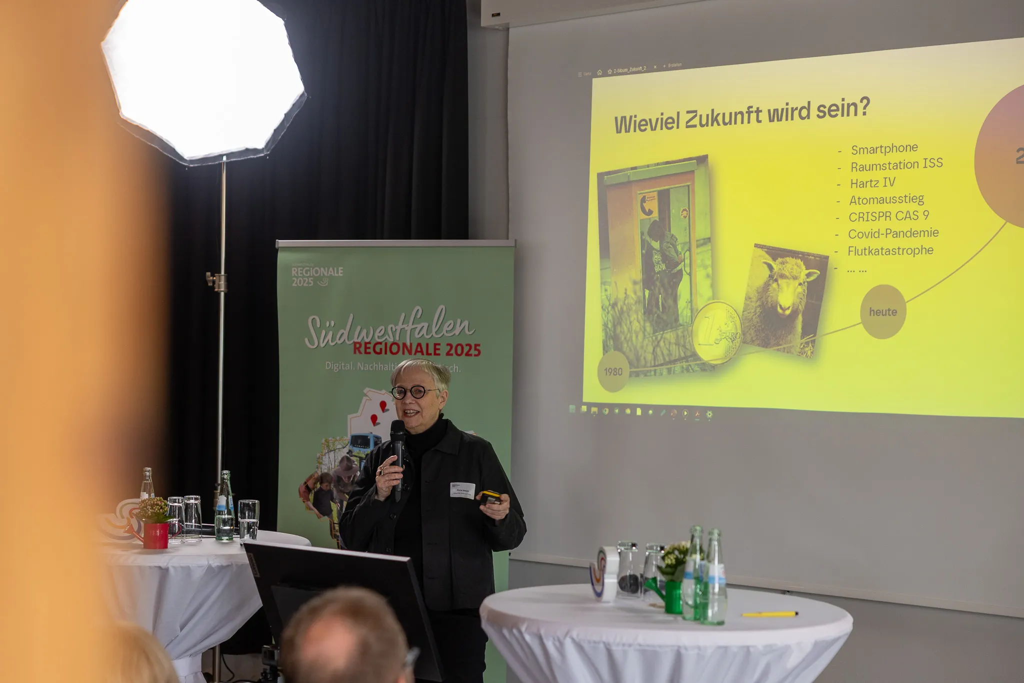 Ein Mann mit Brille hält ein Mikrofon und spricht auf einer Konferenz vor einer Leinwand mit der Aufschrift „Wieviel Zukunft wird sein?“ und einer Grafik, die den Wandel von 1980 bis heute darstellt.