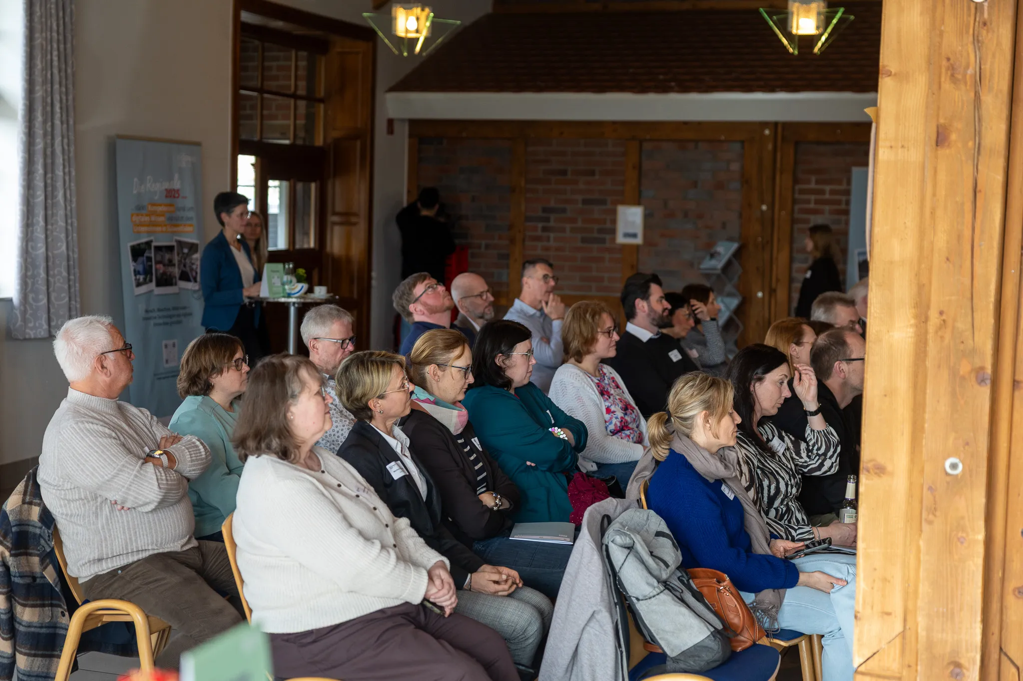 Zuhörer sitzen auf Stühlen in einem hellen Raum und verfolgen eine Präsentation. Im Hintergrund ein Banner mit dem Titel 'Vielfalt Wohnen und Arbeiten'.