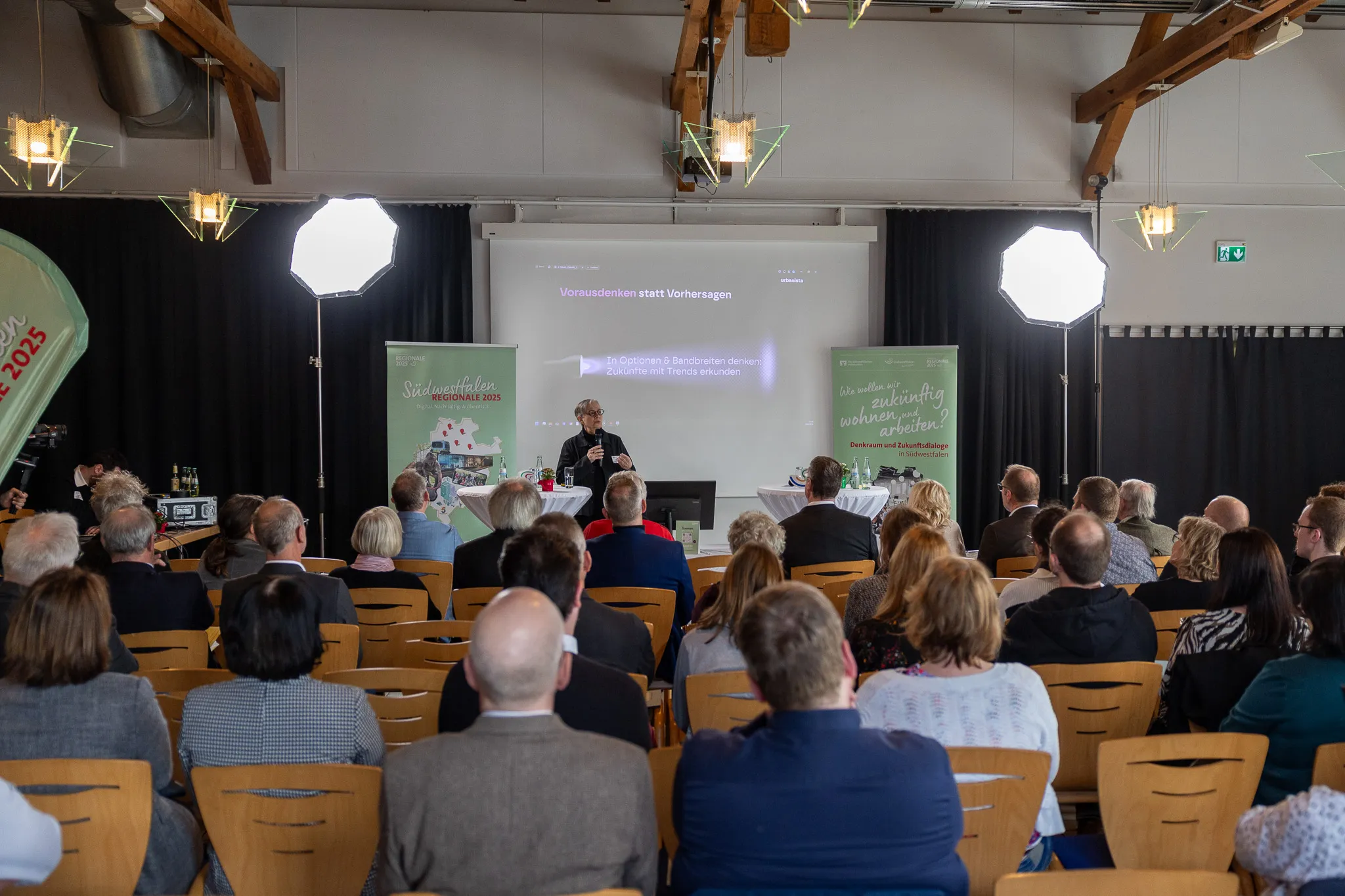 Ein Mann spricht auf einer Konferenz vor einem Publikum. Im Hintergrund eine Leinwand mit der Aufschrift 'Vorausschau statt Vorhersagen' und Logos der 'REGIONALE 2025 Südwestfalen'.