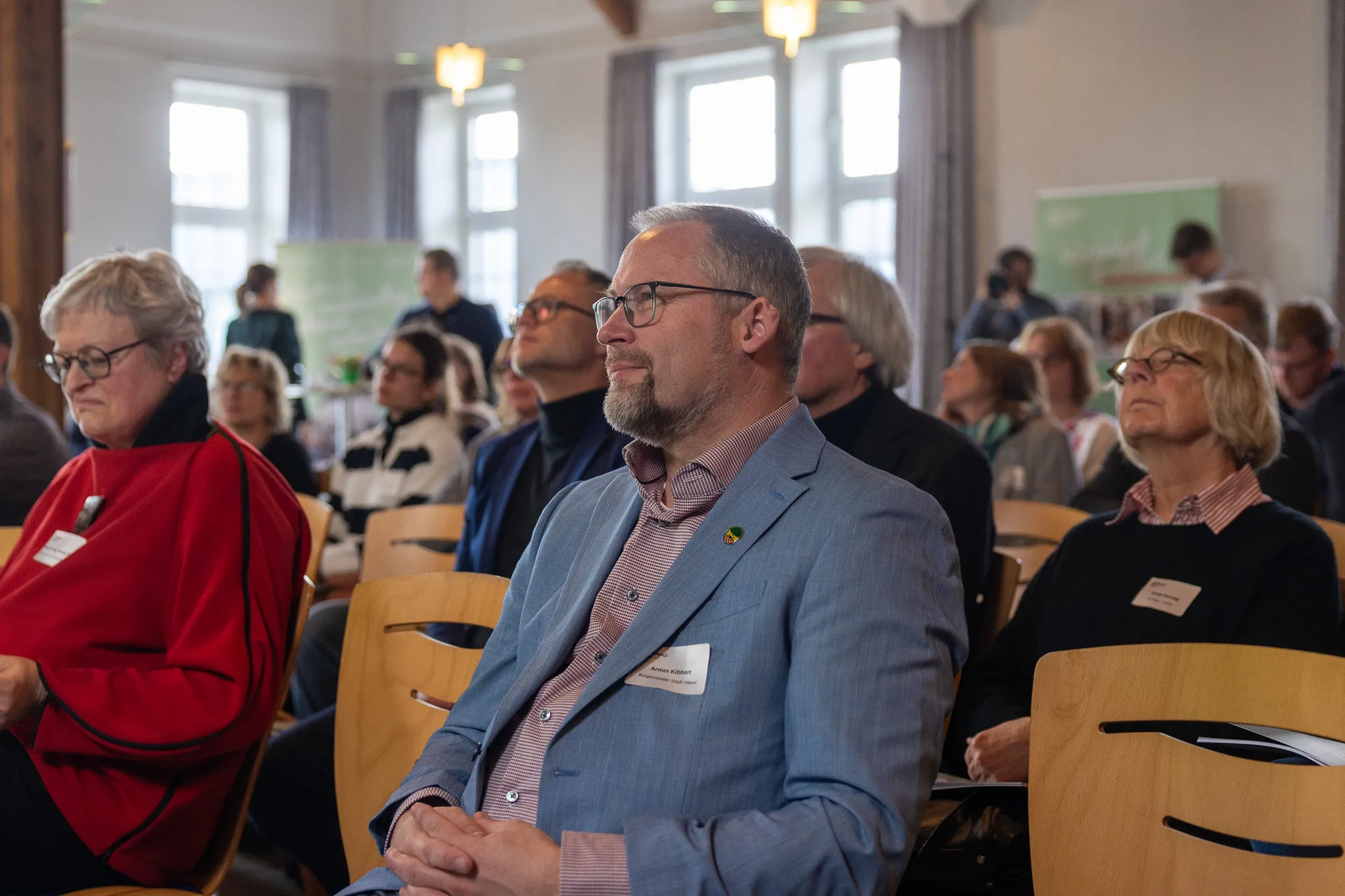 Ein Mann mit Brille im blauen Anzug sitzt auf einer Konferenz und blickt nach vorn. Im Hintergrund weitere Konferenzteilnehmer in einem hellen Raum.