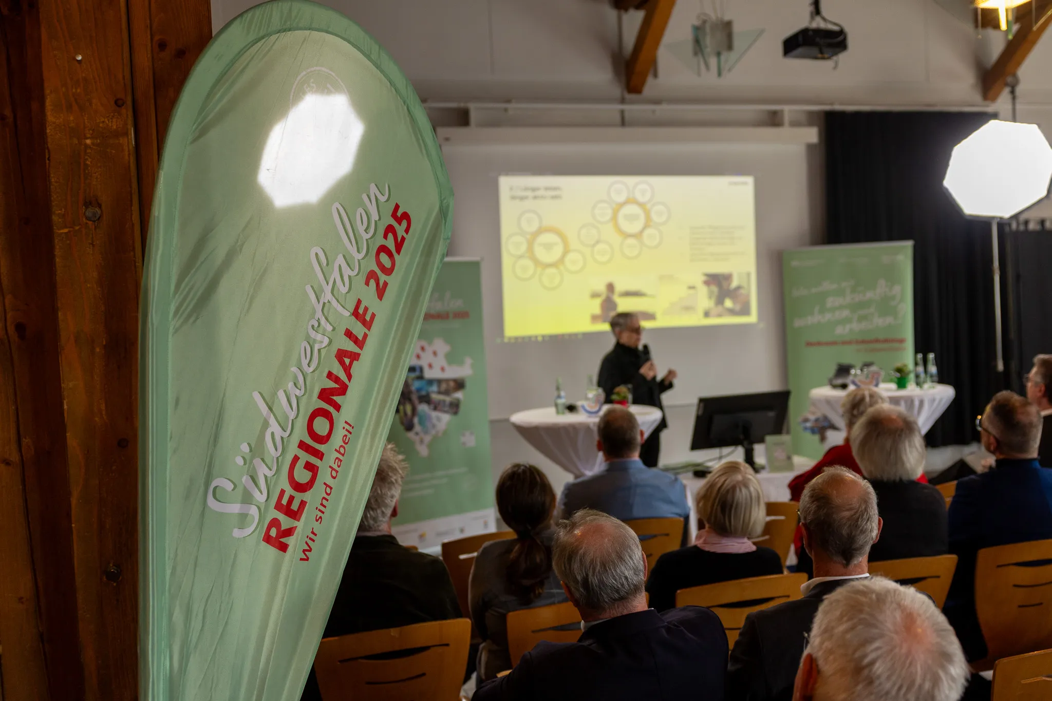 Ein Sprecher steht vor einer Projektion und einem Publikum in einem hellen Raum. Im Vordergrund ein grüner Banner mit der Aufschrift „Südwestfalen REGIONALE 2025 – Wir sind dabei!“