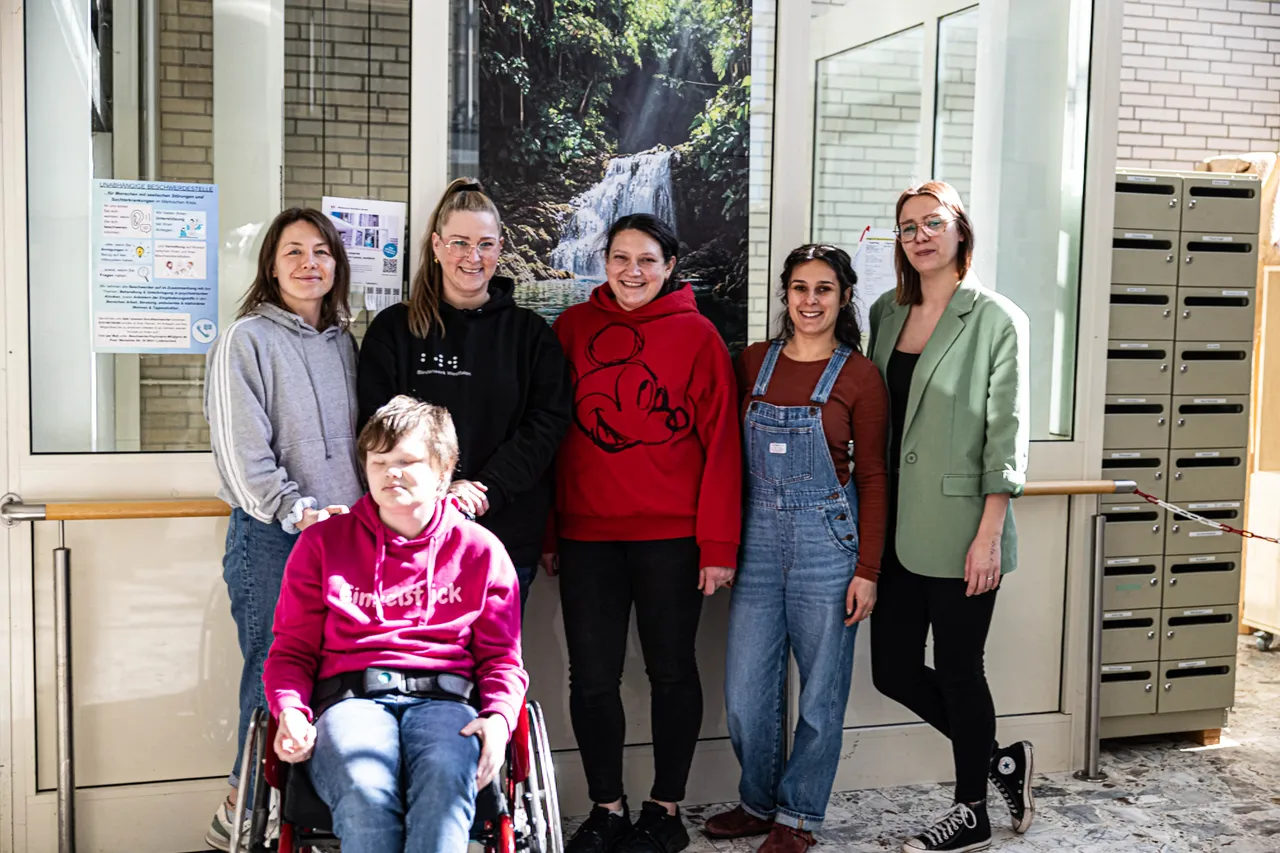 Fünf Frauen und eine Rollstuhlfahrerin stehen vor einem großen Bild eines Wasserfalls und Schließfächern. Sie repräsentieren das Blindenwerk Westfalen und bitten um Spenden für die Reparatur eines defekten Aufzugs.