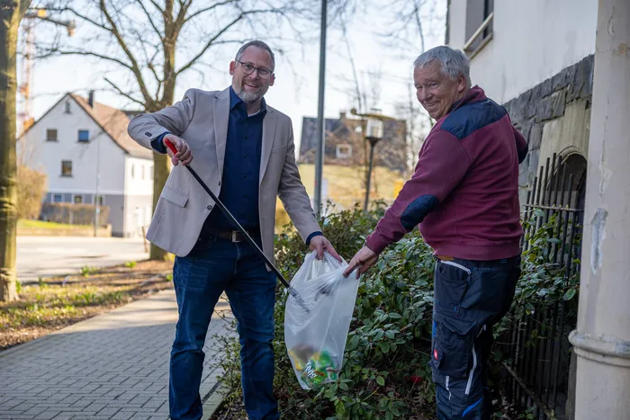Zwei Männer sammeln Müll am Straßenrand. Der eine trägt Anzug, der andere Sportkleidung und beide lächeln in die Kamera.