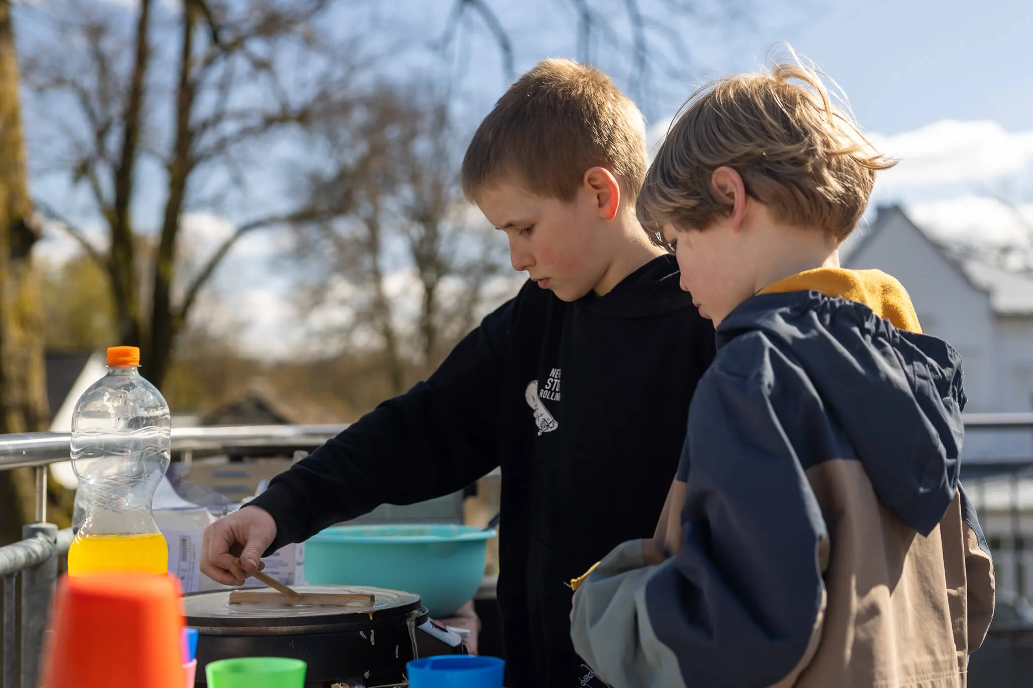 Zwei Jungen bereiten Crêpes auf einem mobilen Grill zu, umgeben von Schüsseln und Zutaten. Im Hintergrund sind Häuser und ein sonniger Himmel zu sehen.