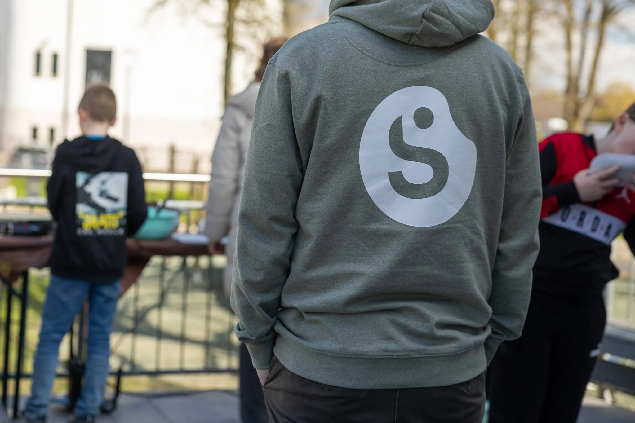 Rückenansicht einer Person mit einem grauen Kapuzenpullover mit großem weißen Kreis und dem Buchstaben S darauf. Im Hintergrund sind Kinder und Erwachsene auf einem Spielplatz zu sehen.