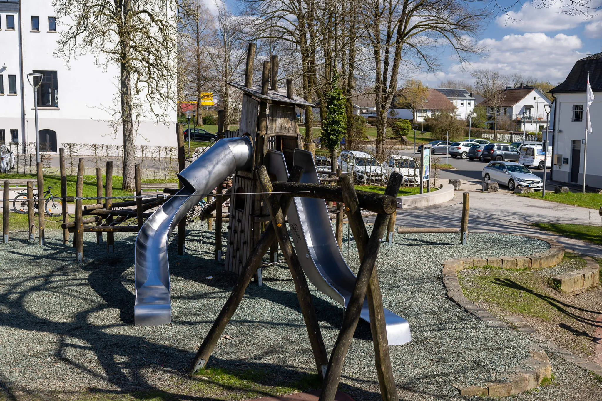 Zwei glänzende Metallrutschen an einem Holzspielturm auf einem Spielplatz mit Holzsägemehl als Fallschutz.