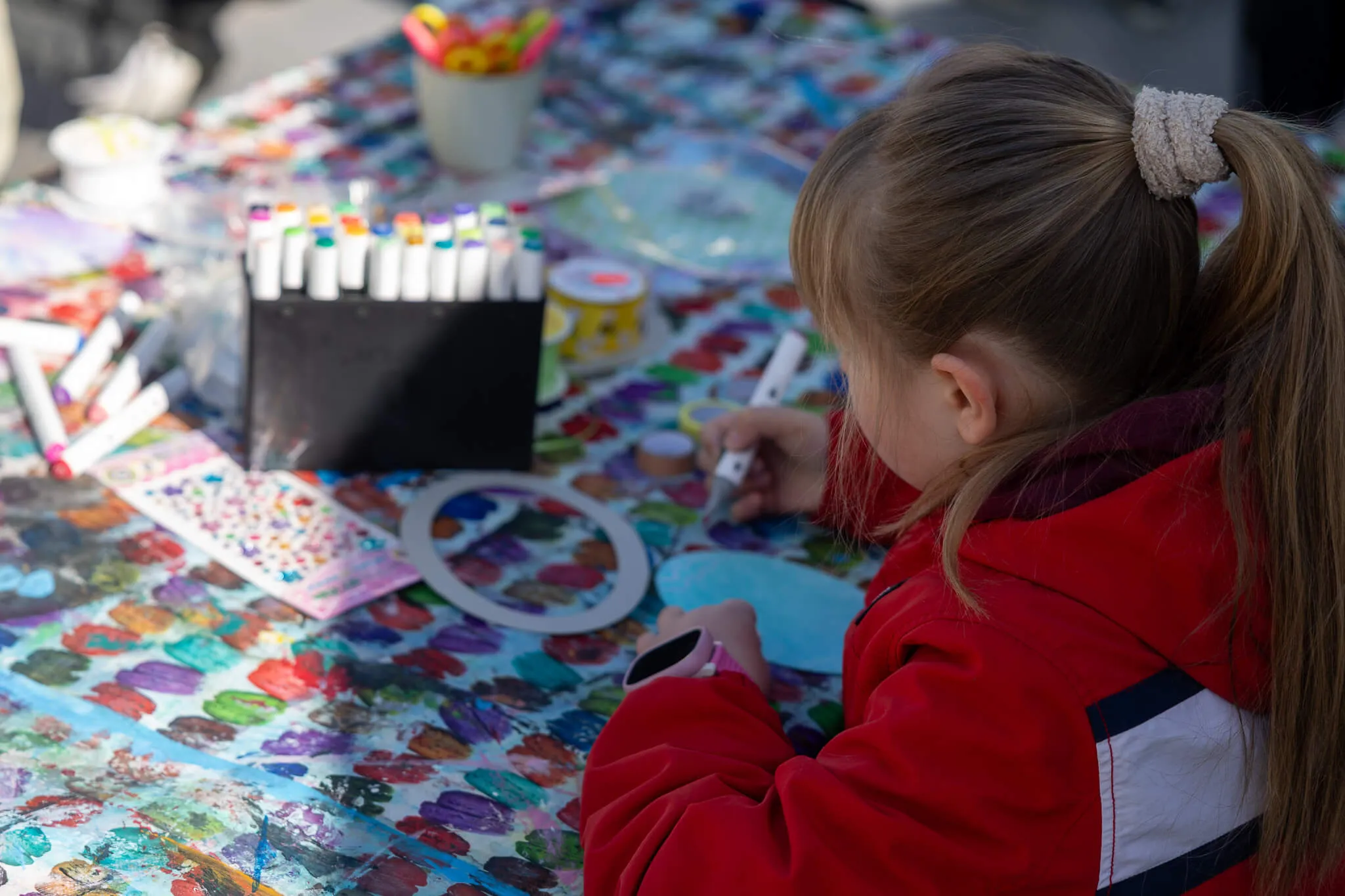 Ein Mädchen mit hochgestecktem Pferdeschwanz bemalt einen Pappteller an einem Tisch voller Bastelmaterialien.