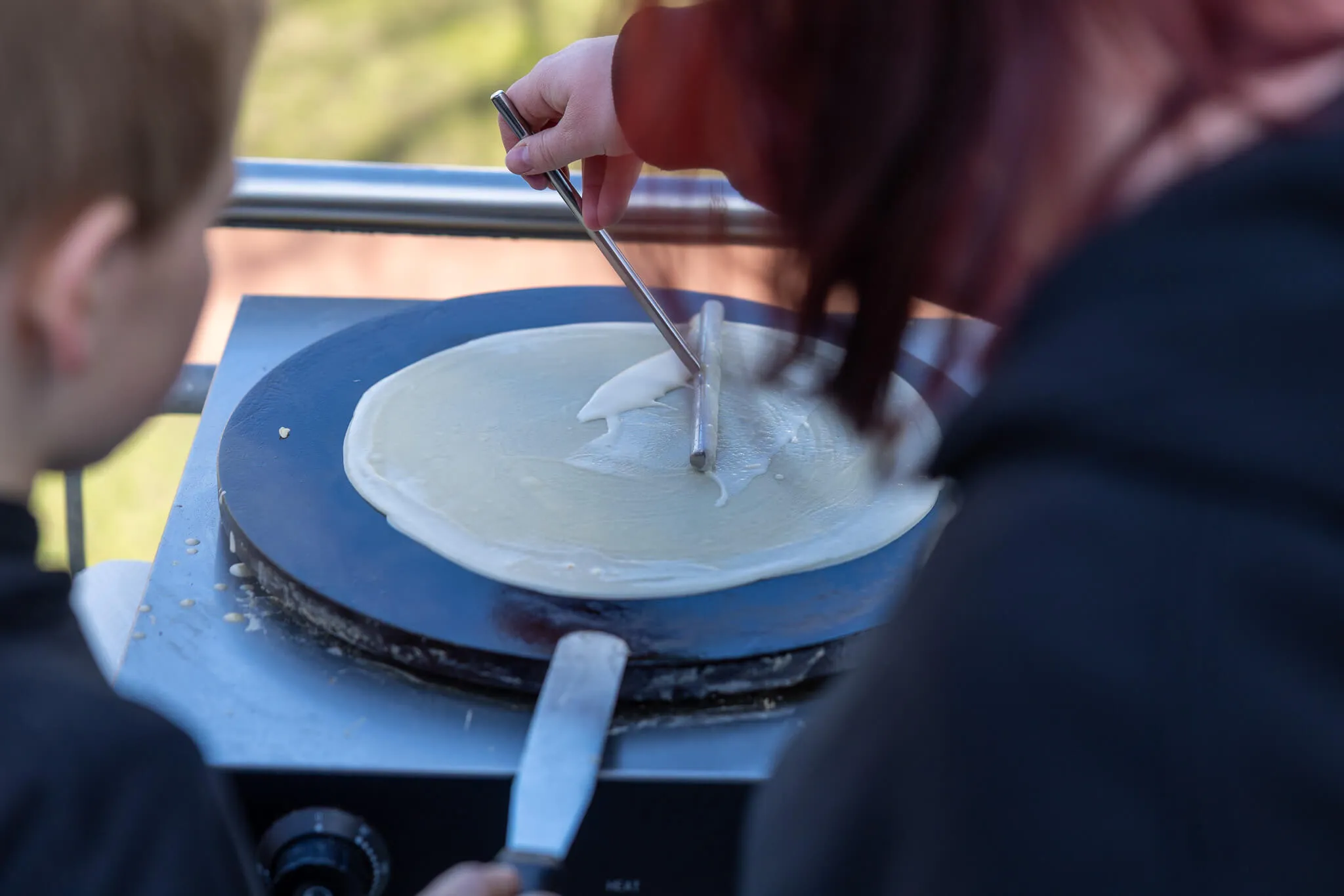Eine Person verteilt mit einem Werkzeug Belag auf einem großen Crêpe, der auf einer heißen Platte zubereitet wird. Im Hintergrund ist ein Kind zu sehen.