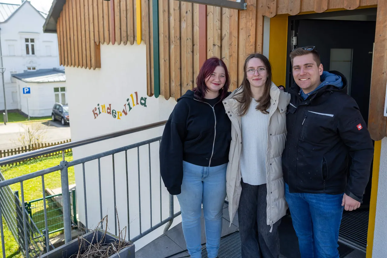 Drei junge Erwachsene stehen vor einem hellen Gebäude mit dem Schriftzug „Kindervilla“. Im Hintergrund ist ein Parkplatz und ein Wohnhaus zu sehen.