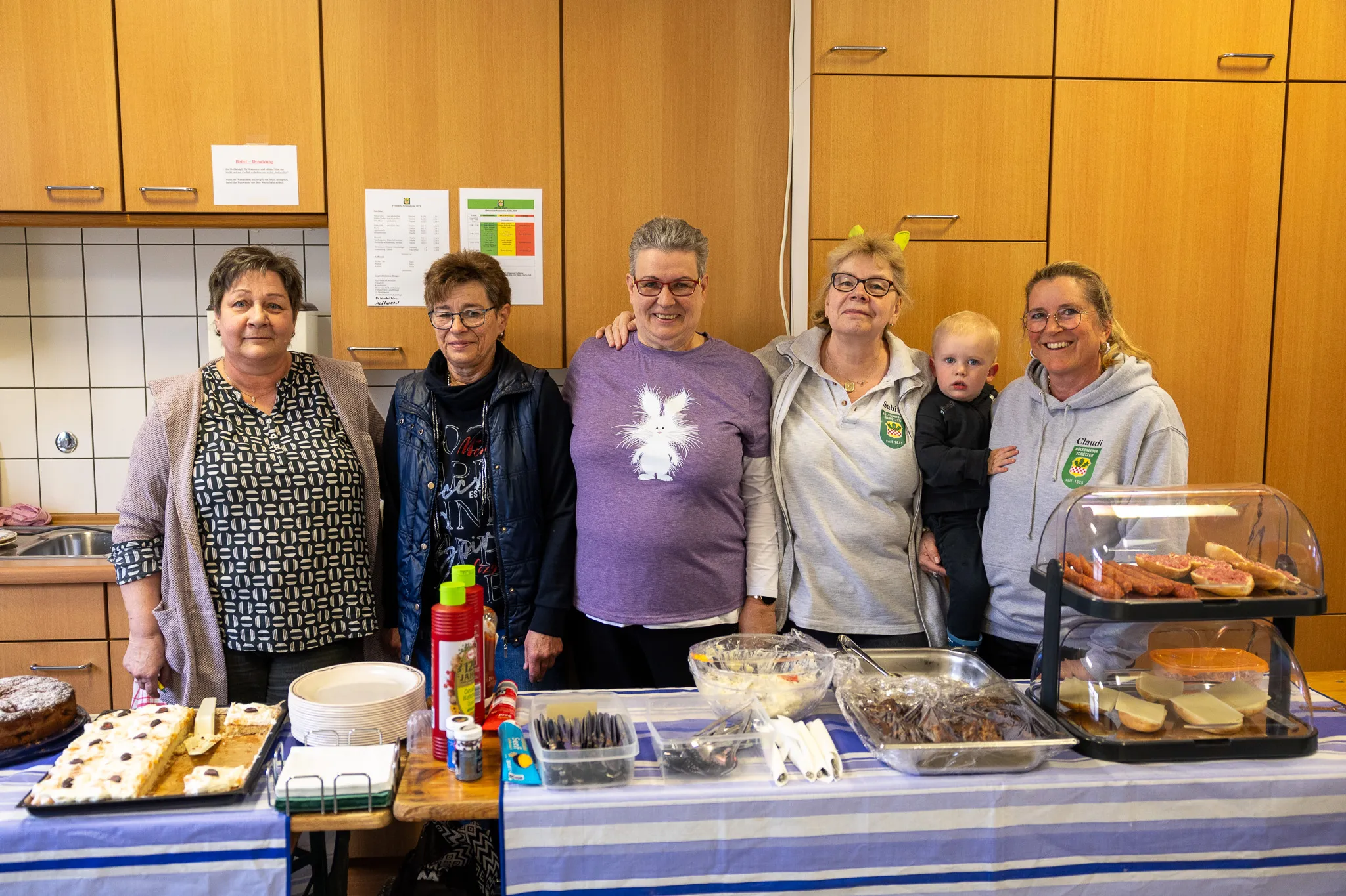 Fünf Frauen und ein Kleinkind stehen lächelnd vor einem mit Speisen gefüllten Tisch. Im Hintergrund hängen Plakate an einer gefliesten Wand.