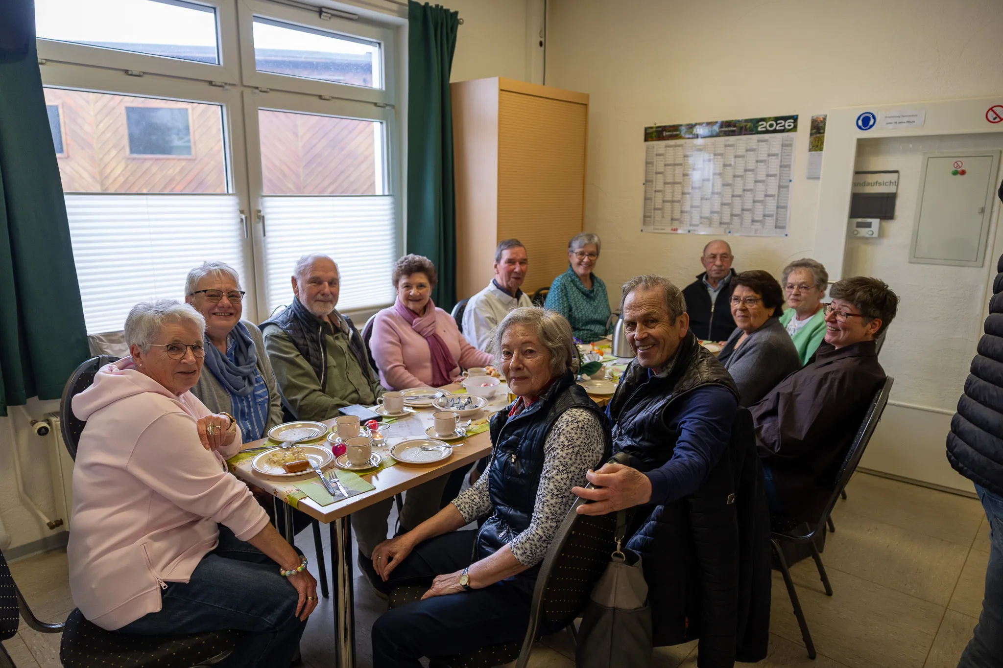Eine Gruppe älterer Menschen sitzt an einem Tisch in einem hellen Raum und isst Kuchen. Im Hintergrund ein Kalender mit dem Jahr 2026.