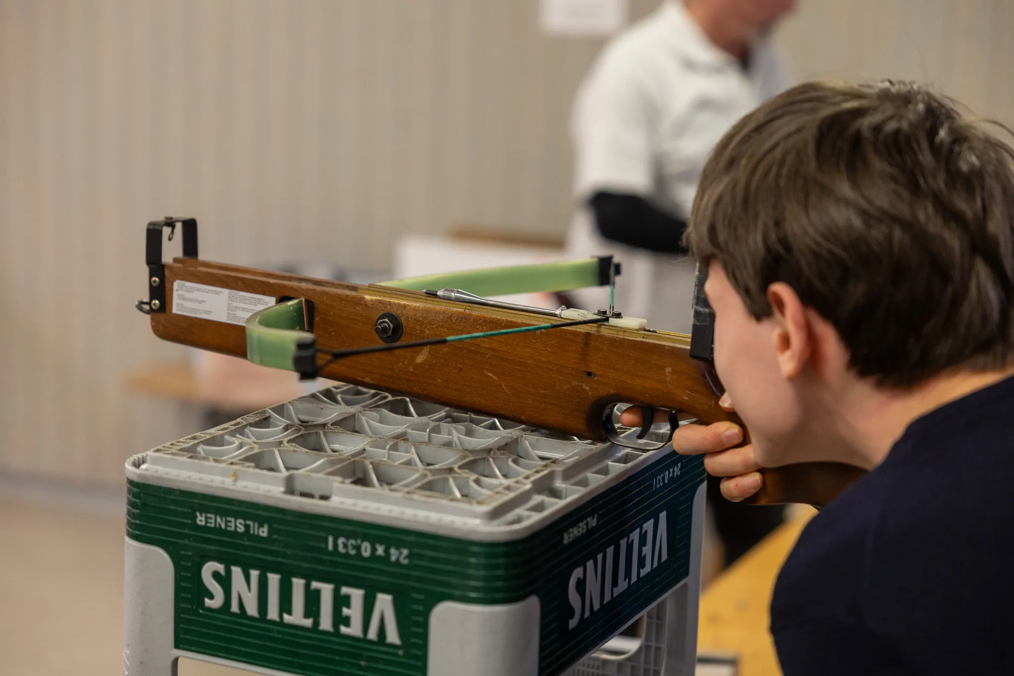 Ein Junge zielt mit einer Armbrust auf eine Zielscheibe. Die Armbrust steht auf Kisten mit dem Aufdruck „Veltins“.