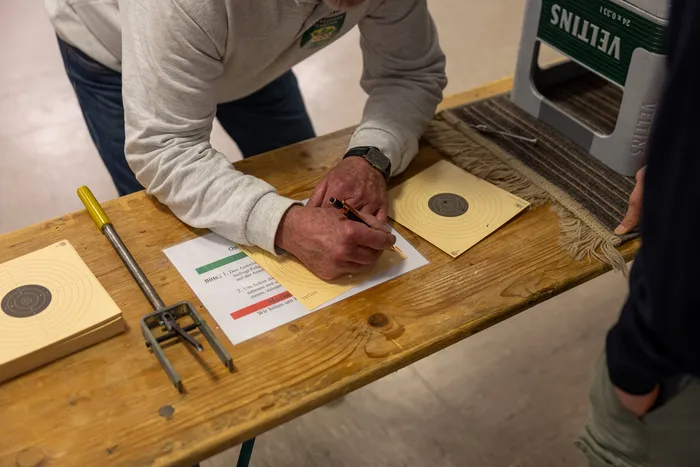 Ein Mann in grauer Jacke schreibt auf ein Blatt Papier, während im Hintergrund weitere Personen und Zielscheiben zu sehen sind. Das Bild zeigt das Ostereierschießen der Hülscheider Schützen.