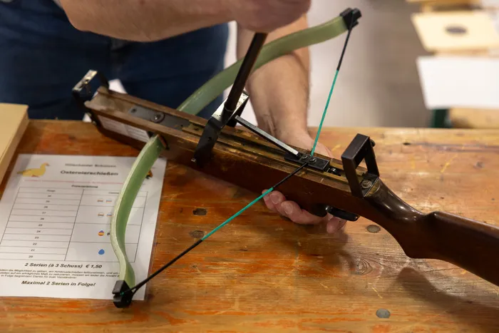 Hände halten eine Armbrust über einem Schießstand mit Zielscheiben in Form von Ostereiern. Im Hintergrund ist ein Punktestand zu sehen.