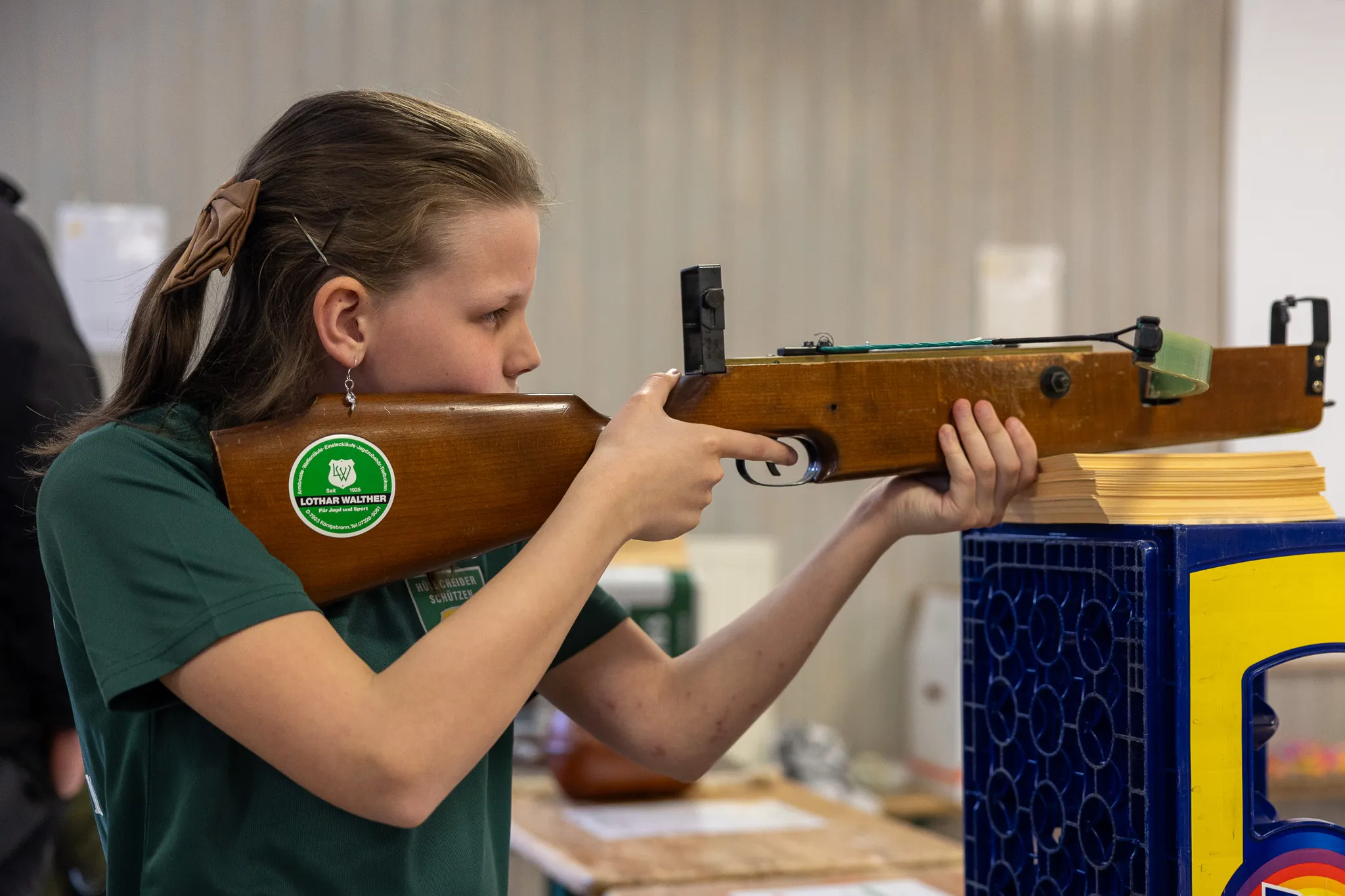 Eine junge Schützin zielt konzentriert mit einem Luftgewehr auf eine Zielscheibe. Sie trägt ein grünes T-Shirt mit dem Aufdruck „Schützen“ und hat ihr Haar zu einem Zopf gebunden.