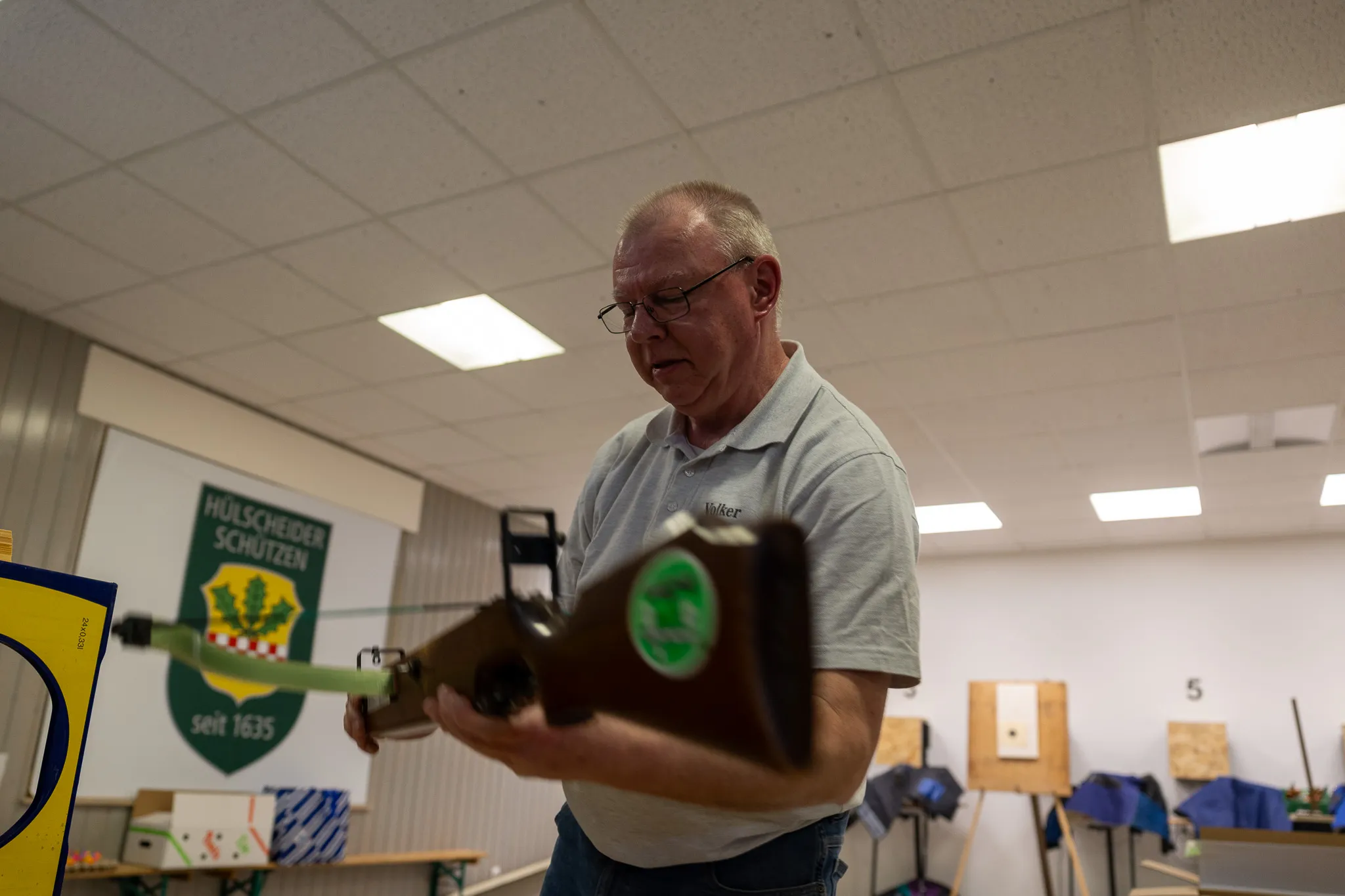 Ein älterer Mann mit Brille und blauem Hemd hält einen Luftgewehr vor sich und justiert es. Im Hintergrund ein Banner mit dem Aufdruck „Hülscheider Schützen seit 1635“.