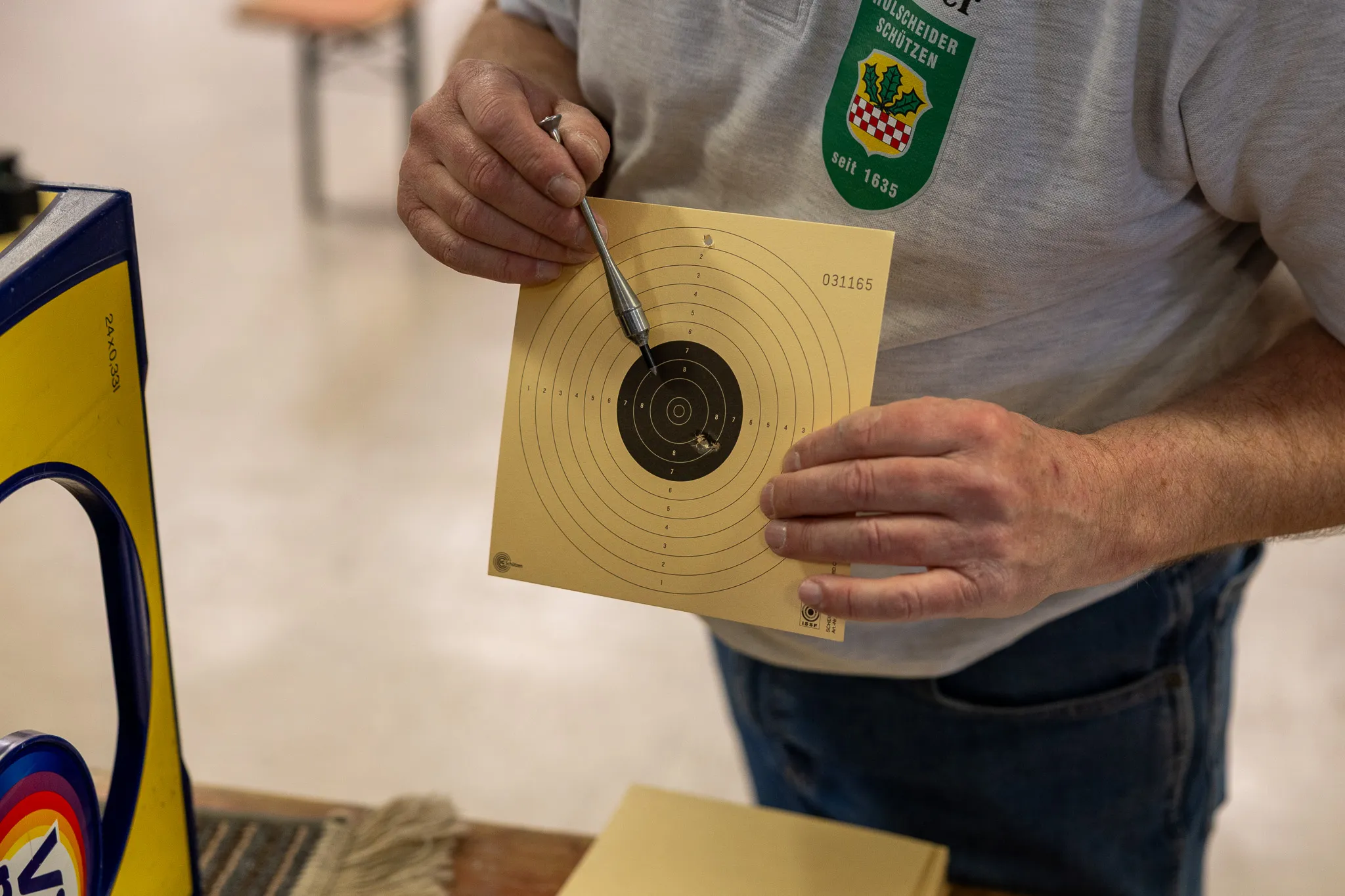 Eine Person hält eine Zielscheibe mit mehreren Schusslöchern in der Hand. Im Hintergrund ist ein Teil einer Zielanlage und eine Person mit einem Abzeichen zu sehen.