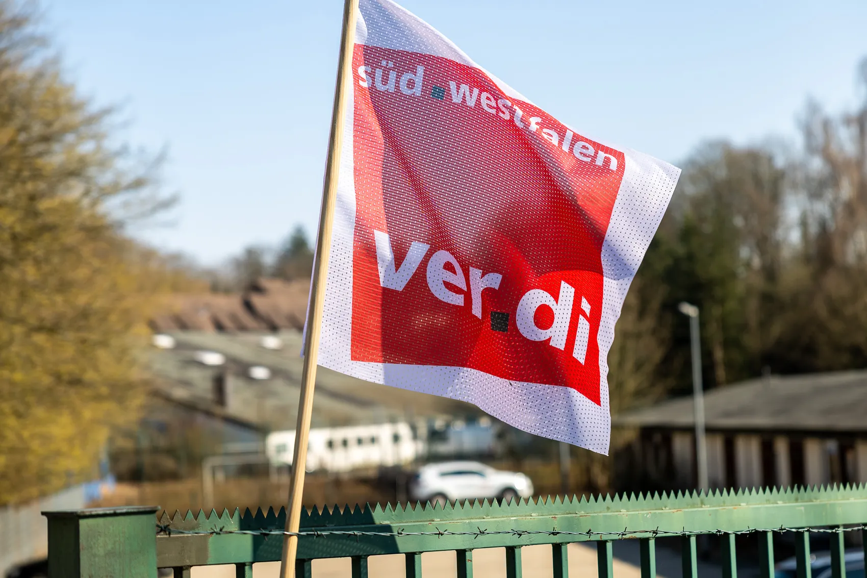 Rot-weiße Verdi-Flagge mit dem Schriftzug „ver.di süd westfalen“ weht vor einem Stacheldrahtzaun und Gebäuden im Hintergrund.
