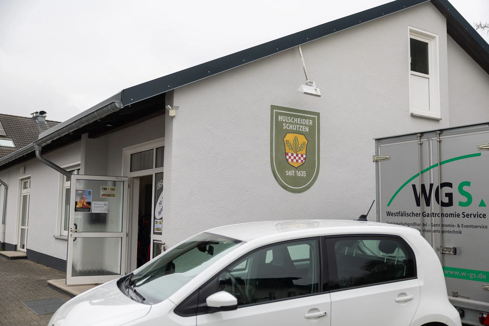 Ein hellgraues Gebäude mit dem Wappen der Hülscheider Schützen und dem Schriftzug „seit 1635“. Davor steht ein schwarzer PKW und ein weißer Transporter der Westfälischen Gastronomie Service WGS.